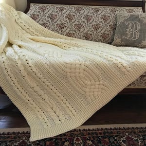 May include: A cream-colored crocheted throw blanket with a cable knit pattern is draped over a couch. The blanket has a delicate, lacy texture and is made with a soft yarn.