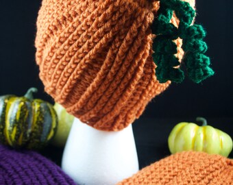 May include: A crocheted pumpkin hat with a green stem and leaves. The hat is displayed on a white mannequin. There are three small pumpkins in the background.
