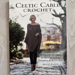 May include: Book cover featuring a woman wearing a brown crocheted poncho and black pants. The title of the book is "Celtic Cable Crochet" and the subtitle is "18 Crochet Patterns for Modern Cabled Garments & Accessories". The author is Bonnie Barker.