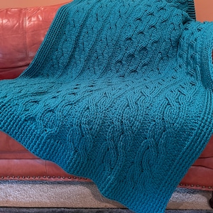 May include: A teal blue crocheted blanket with a cable knit pattern. The blanket is draped over a brown leather couch.