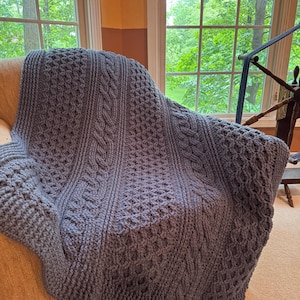 Op de afbeelding: Een blauwe gehaakte deken met ingewikkelde kabel- en textuursteekpatronen over een stoel gedrapeerd. De deken heeft verschillende ontwerpen, waaronder verticale kabelrijen en ruitvormig openwerk, waardoor een gezellig en decoratief textiel ontstaat.