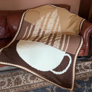May include: A handmade crochet blanket featuring a coffee cup design. The blanket is primarily brown, beige, and white, with a detailed border. The top portion has a textured design. The blanket is draped over a brown leather couch.