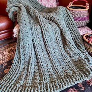 Op de afbeelding: Een saliegroene, dikke gebreide deken over een stoel. De deken heeft een gestructureerd, geribbeld patroon met een zachte, pluche uitstraling. De stoel heeft een bloemenpatroon op de rug en staat op een patroonkleed.