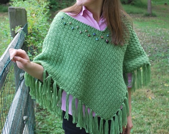 Op de afbeelding: Een groene gebreide poncho met franjes en kleurrijke kralen rond de halslijn.