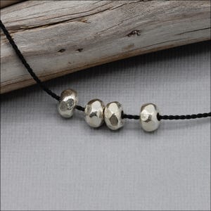 Pu&ograve; includere: Una collana con cordino nero caratterizzata da quattro perline color argento con una finitura martellata e testurizzata. Le perline sono distanziate uniformemente lungo il sottile cordino nero intrecciato, creando un design semplice e pulito.