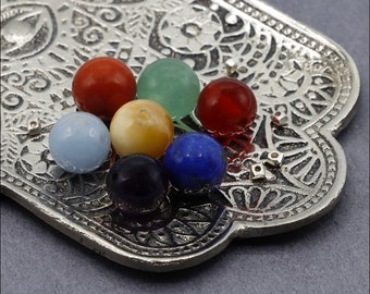 May include: A silver Hamsa hand charm with seven colorful gemstones in the center. The gemstones are red, green, cream, blue, purple, light blue, and orange.