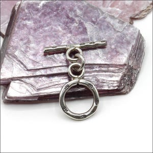 May include: A silver toggle clasp with a circular ring and a bar. The ring has decorative floral patterns around the edge. The bar has similar floral patterns. The clasp is connected by three interlocking rings. The background is a layered, purple-toned stone.