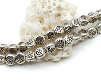 May include: A strand of metallic silver-toned beads, each featuring a square shape with rounded edges and a stamped heart design on the surface. The beads are arranged in two rows, showcasing a consistent polished finish and a repeating heart motif.