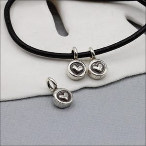 May include: Three small, circular silver-toned metal charms, each featuring a raised heart design in the center. Two of the charms are threaded onto a thin, black cord, while the third charm is positioned separately below them on the surface.
