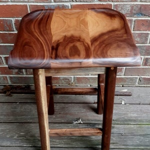May include: A wooden stool with a curved top and a dark brown finish. The stool has a simple design with three legs and a single crossbar.