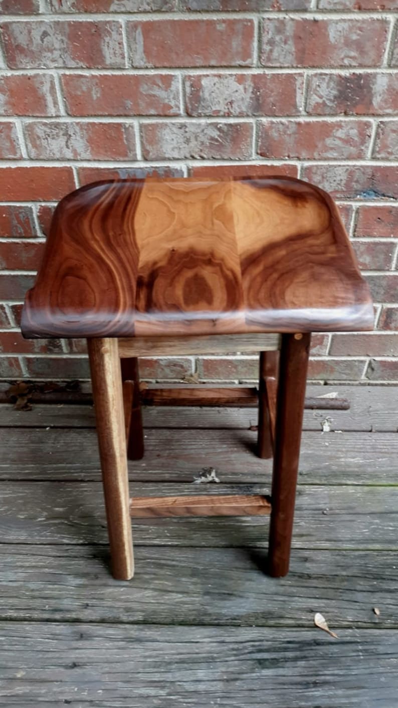 May include: A wooden stool with a curved top and a dark brown finish. The stool has a simple design with three legs and a single crossbar.
