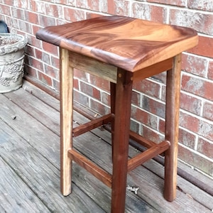 May include: A wooden stool with a curved top and a natural wood finish. The stool has a simple design with four legs and a single crossbar.