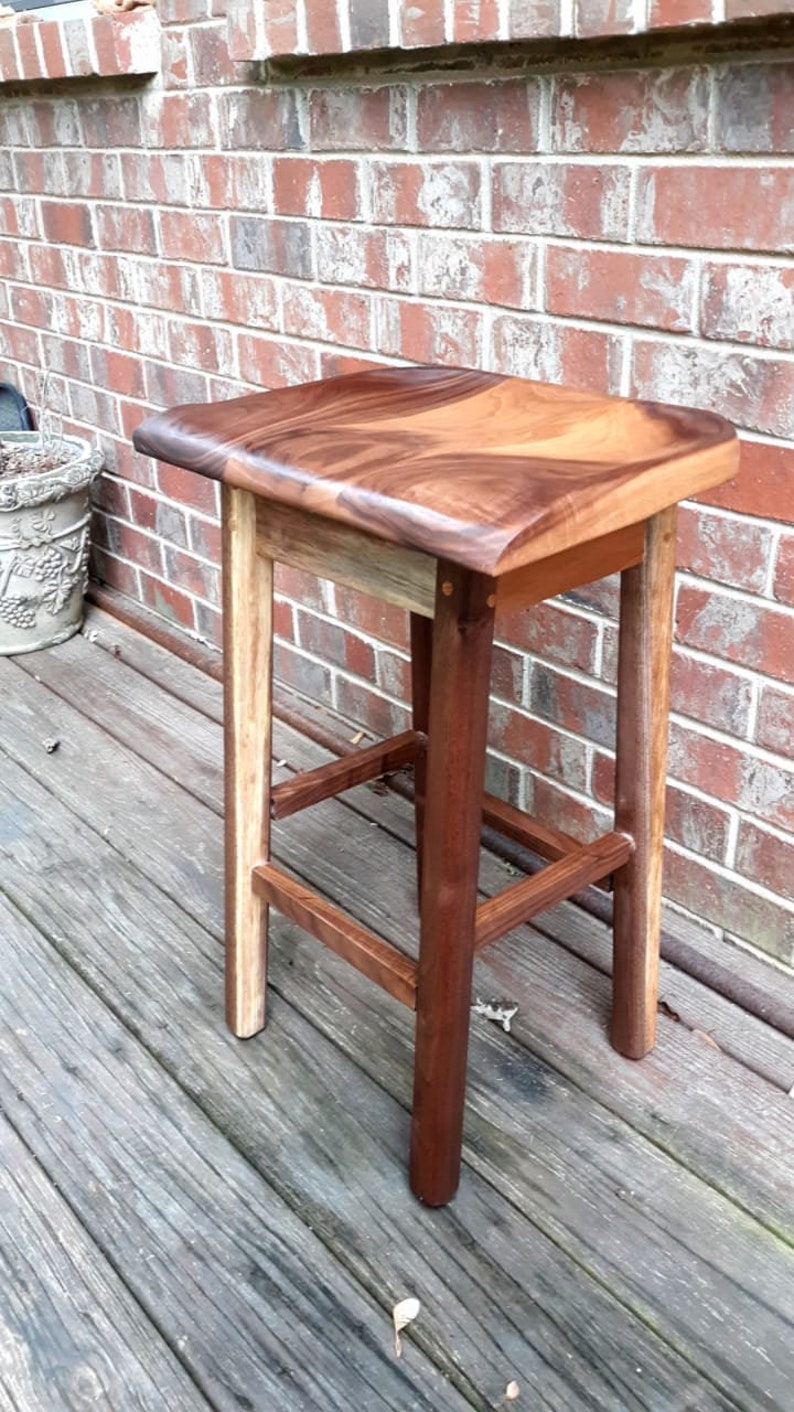 May include: A wooden stool with a curved top and a natural wood finish. The stool has a simple design with four legs and a single crossbar.