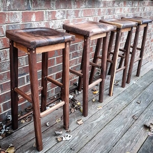 May include: Five wooden bar stools with a dark brown finish and a natural wood seat. The stools are lined up in a row against a brick wall.