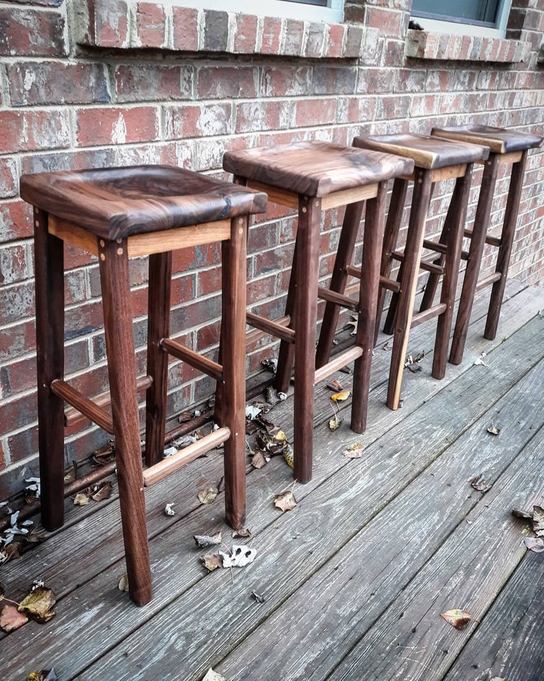 May include: Five wooden bar stools with a dark brown finish and a natural wood seat. The stools are lined up in a row against a brick wall.