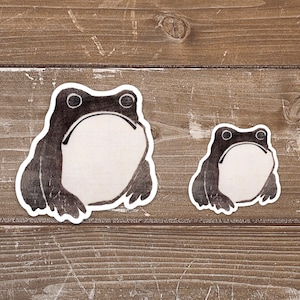 May include: Two black and white frog stickers on a wood background. The larger frog is on the left and the smaller frog is on the right.