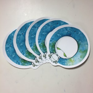 May include: Five white circular stickers with a blue and green watercolor galaxy design. Each sticker features a white cartoon alien with green eyes looking out from the galaxy.