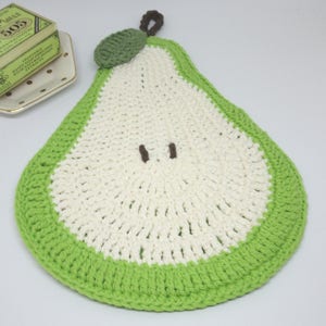 May include: A crocheted pear-shaped pot holder with a green border and a green leaf on top. The pot holder is white in the center with two brown stitches for eyes.