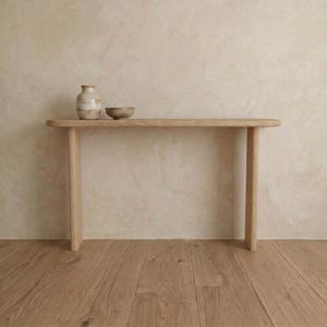 May include: A light-colored wooden console table with a rounded top and two legs. A small, beige ceramic vase and a matching bowl sit on the table. The background is a textured, off-white wall, and the floor is made of light-colored wood.