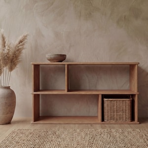 May include: A light brown wooden shelf unit with open compartments and a woven basket. A decorative vase with dried plants and a wooden bowl are also present. The shelf is made of light-colored wood and has a simple, modern design.