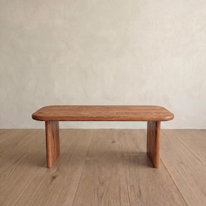 May include: A low, rectangular coffee table made of wood, featuring rounded corners and a natural wood grain. The table, with its warm reddish-brown colour, is supported by two U-shaped legs. It sits on a light wooden floor. The table measures approximately 120 cm in length.