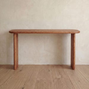 May include: A wooden console table with a natural finish. The table has a rectangular top with rounded corners and two rectangular legs. The wood grain is visible. The table is set against a neutral background. The table's dimensions are in centimetres.
