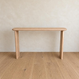 May include: A light brown wooden console table with a long, rectangular top and two vertical slab legs. The table features a smooth, natural wood grain finish with rounded edges on the top surface, creating a clean and simple structural design.