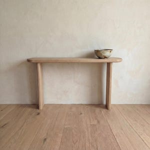 May include: A light-colored wooden console table with a rounded top and two legs. A small, textured bowl with brown and beige tones sits on the table. The table is set against a neutral-toned wall and hardwood floor.