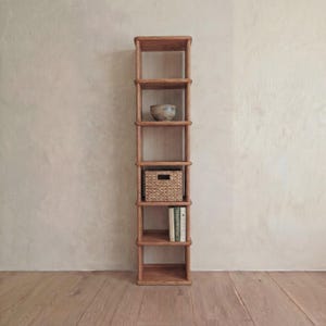 May include: A tall wooden bookshelf with six shelves. A decorative bowl, a woven basket, and two books are displayed on the shelves. The bookshelf is made of light brown wood and has a simple, modern design.