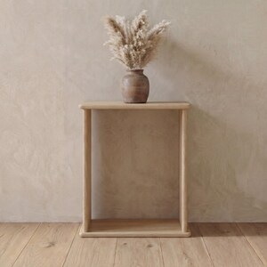 May include: A light-coloured wooden console table with a rectangular top and two vertical side supports. A dark brown ceramic vase sits on the table, containing a large, fluffy arrangement of dried beige pampas grass plumes.