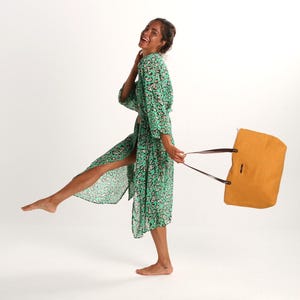 May include: A woman in a green floral print robe holds a mustard yellow tote bag with brown straps. She is smiling and walking with one leg raised.