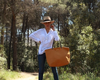 Pode incluir: Uma mulher com uma camisa branca, jeans azuis e um chapéu de palha, segurando uma grande bolsa tote marrom com alças pretas. A bolsa tem formato retangular e um detalhe dourado. O fundo mostra uma floresta.