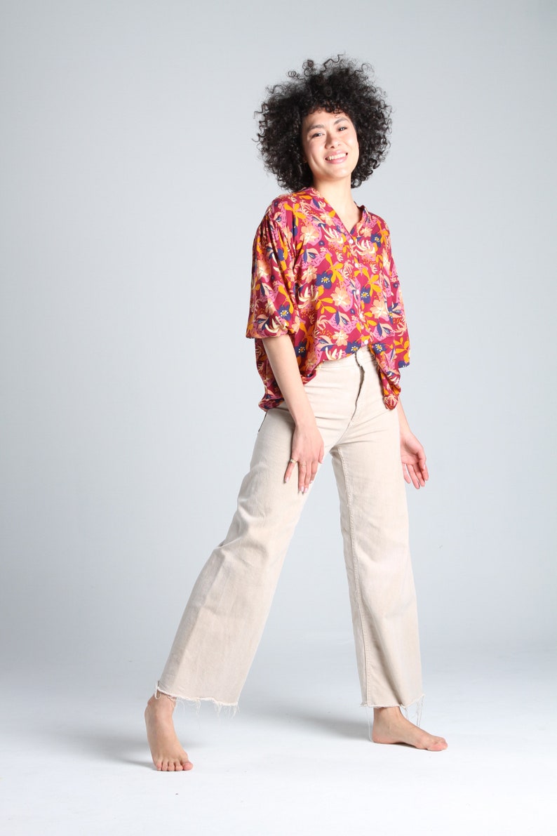 May include: A woman with curly brown hair is wearing a red, yellow, and blue floral print button-down shirt and beige wide-leg pants. She is smiling and looking at the camera.
