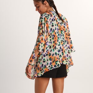 Pode incluir: Uma sa&iacute;da de praia leve e solta em estilo quimono com um padr&atilde;o de leopardo multicolorido vibrante em tons de laranja, roxo e azul-petr&oacute;leo sobre um fundo branco, com mangas largas e abertas e um tecido fluido e semitransparente.
