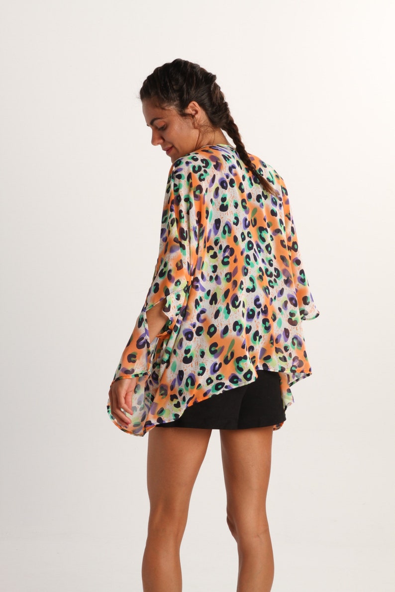Pode incluir: Uma sa&iacute;da de praia leve e solta em estilo quimono com um padr&atilde;o de leopardo multicolorido vibrante em tons de laranja, roxo e azul-petr&oacute;leo sobre um fundo branco, com mangas largas e abertas e um tecido fluido e semitransparente.