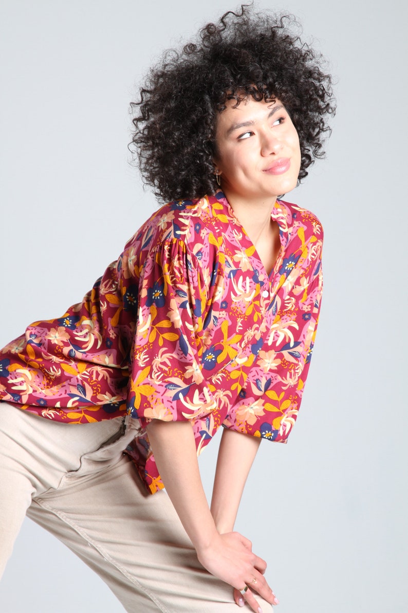 May include: A woman with curly dark hair is wearing a red, pink, yellow, and blue floral print blouse with a button-up collar. She is wearing beige pants and is posing with her hands on her knees.