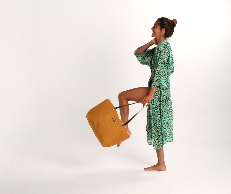 May include: A woman in a green and white floral print robe holds a brown tote bag. The bag has a brown leather handle and a gold metal clasp.