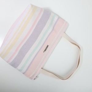 May include: A striped tote bag with a light pink, white, yellow, and purple color scheme. The bag has a single, long, cream-colored handle.
