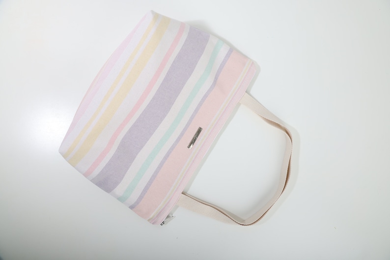 May include: A striped tote bag with a light pink, white, yellow, and purple color scheme. The bag has a single, long, cream-colored handle.