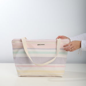 May include: A striped tote bag with a cream canvas strap. The bag is made of a soft, woven fabric and has a zipper closure. The stripes are in shades of pink, yellow, green, and purple.