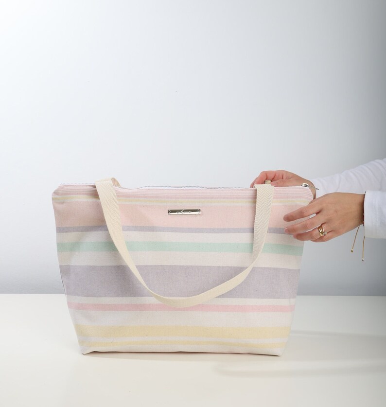 May include: A striped tote bag with a cream canvas strap. The bag is made of a soft, woven fabric and has a zipper closure. The stripes are in shades of pink, yellow, green, and purple.
