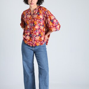May include: A woman wearing a red, yellow, and blue floral print button-up shirt with a relaxed fit and blue denim jeans.
