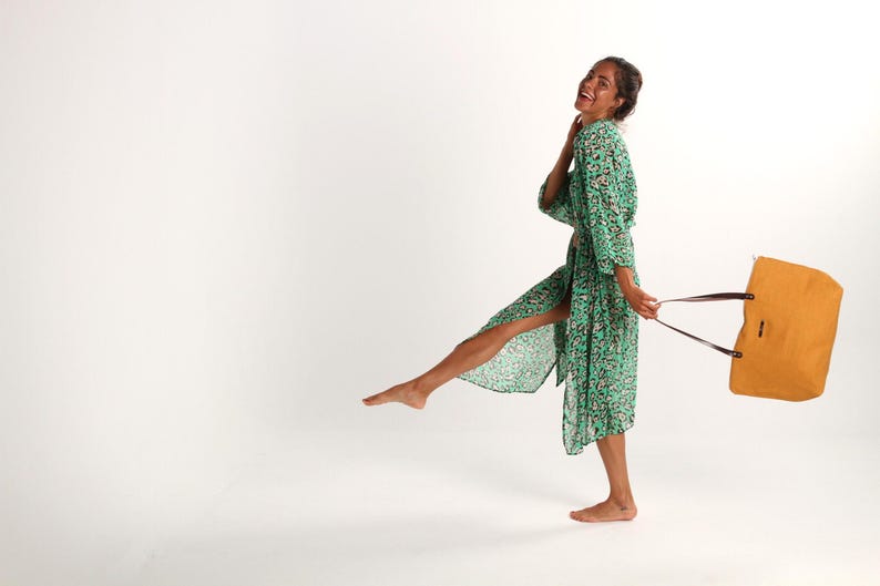 May include: A woman in a green floral print robe holds a mustard yellow tote bag with brown straps. She is smiling and walking with one leg raised.