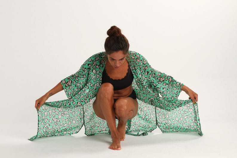 May include: A woman wearing a green and black leopard print kimono with a black bra underneath. The kimono is open and flowing, and the woman is sitting with her legs crossed.