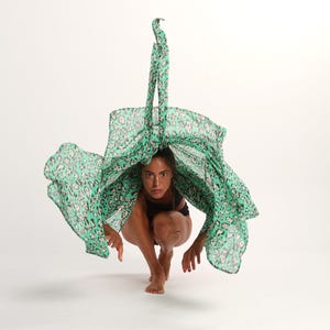 May include: A woman in a black leotard is crouching with a green and black patterned scarf draped over her head. The scarf is flowing in the air.