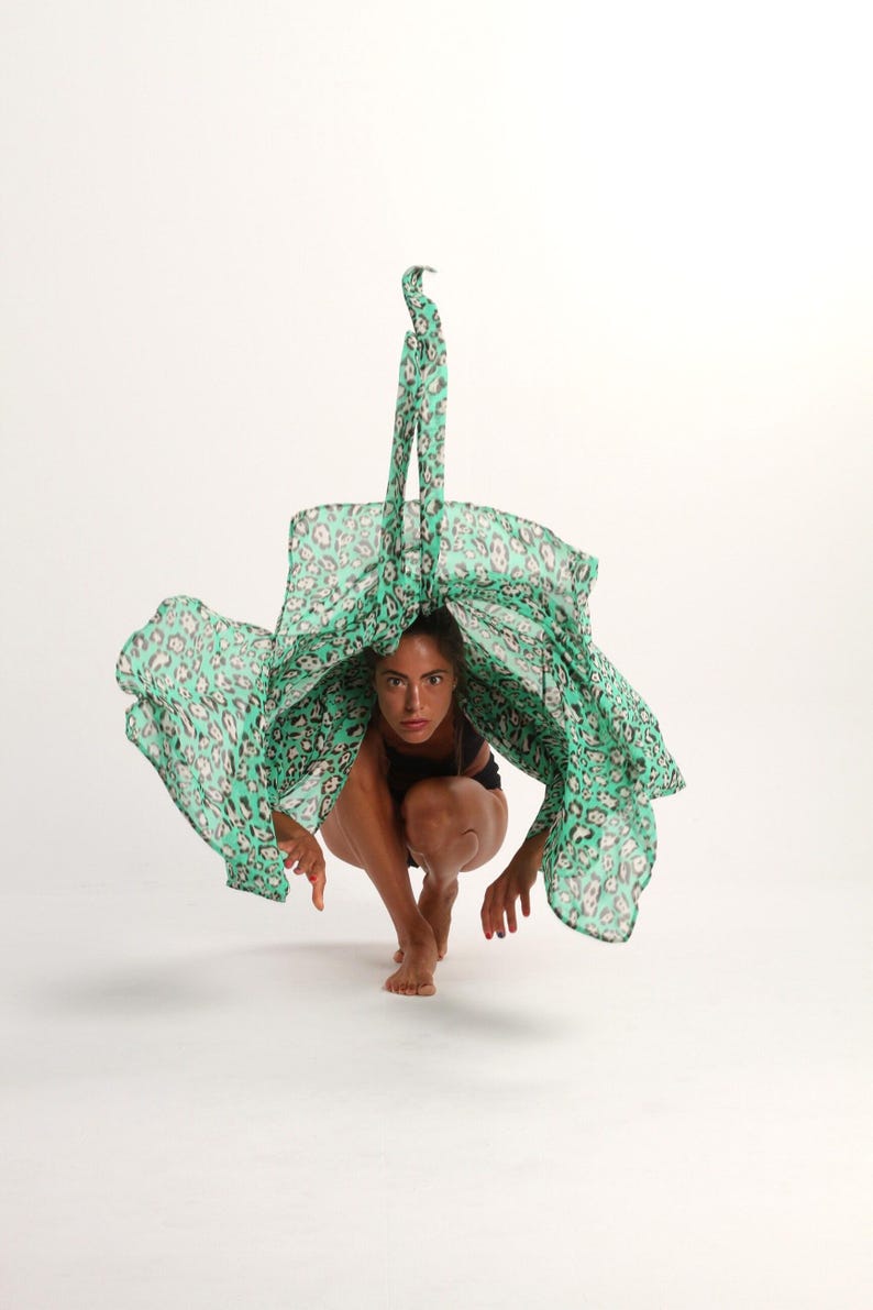 May include: A woman in a black leotard is crouching with a green and black patterned scarf draped over her head. The scarf is flowing in the air.