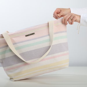 May include: A striped tote bag with a zipper closure and a cream-colored strap. The bag is made of a soft, woven fabric and has a light pink, yellow, green, and purple color scheme.
