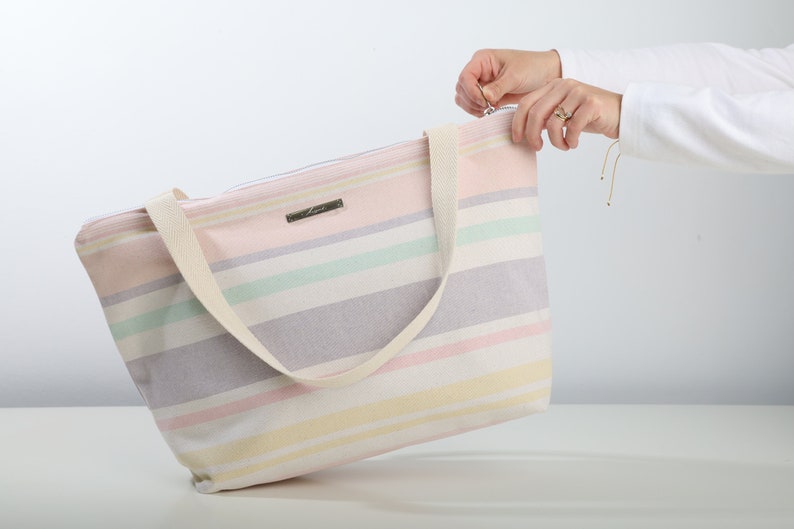 May include: A striped tote bag with a zipper closure and a cream-colored strap. The bag is made of a soft, woven fabric and has a light pink, yellow, green, and purple color scheme.