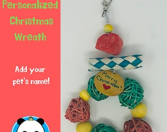 May include: A personalized Christmas wreath for pets, made of natural materials. It features a red, white, and blue striped paper tube, a red sponge, a green and brown woven ball, a yellow bead, and a small wooden tag with the text "Merry Christmas!" written on it. The wreath is attached to a silver clip.