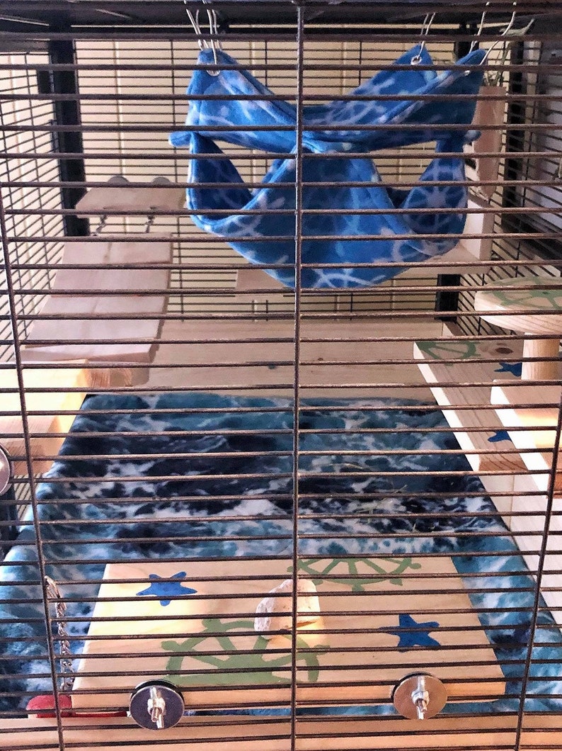 May include: A blue and white patterned fabric hammock hangs inside a cage with wooden platforms and a blue and white patterned fleece bedding.